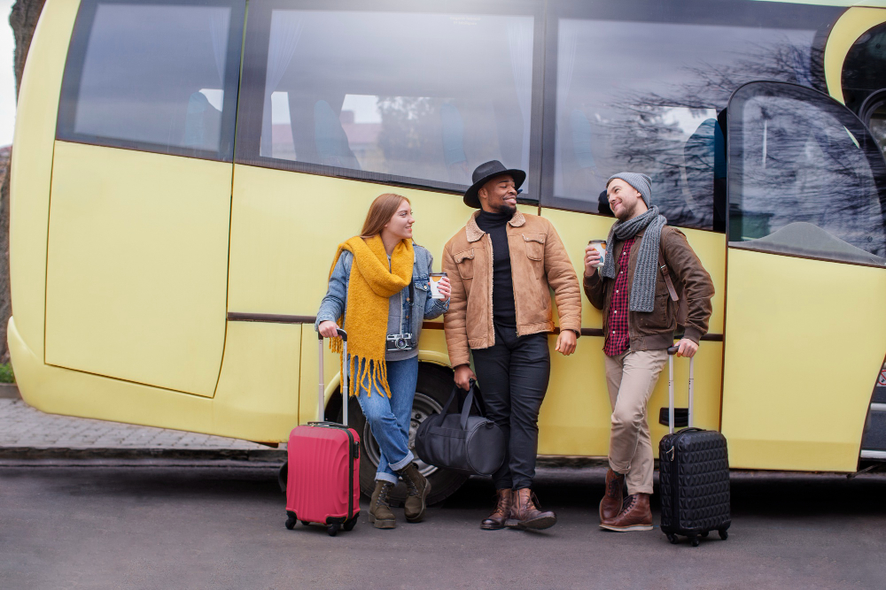 Three friends with rolling luggage stand by a yellow bus, chatting and holding drinks on a city street.