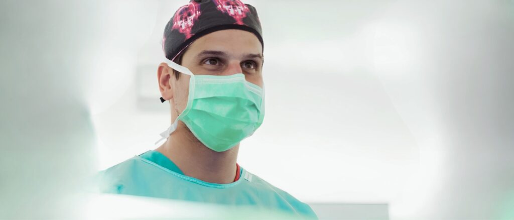 Surgeon wearing a green surgical mask and cap in a bright clinical setting.