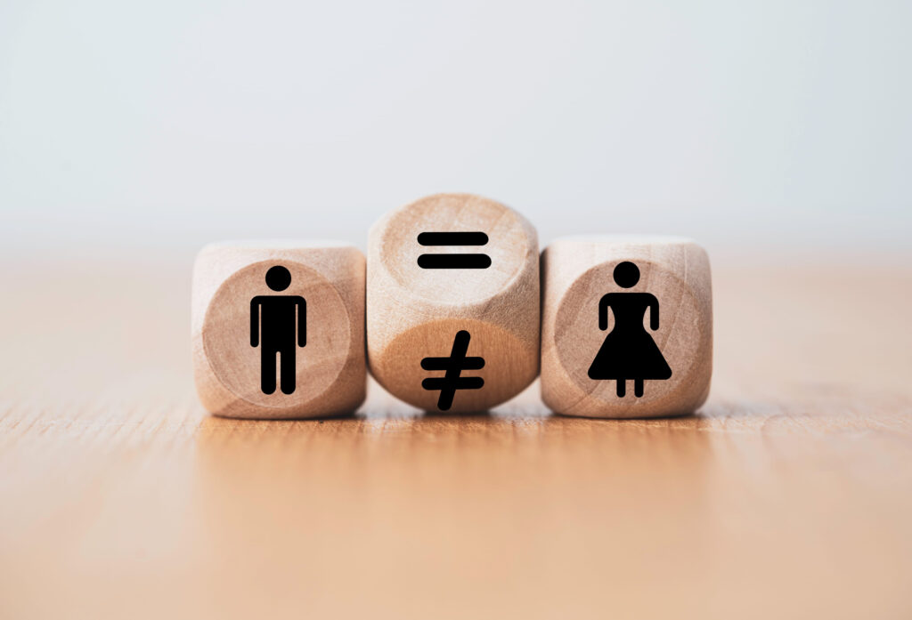 Wooden blocks show a man icon on the left, a middle block with equals and not-equals symbols, and a woman icon on the right, symbolizing gender equality vs. inequality.