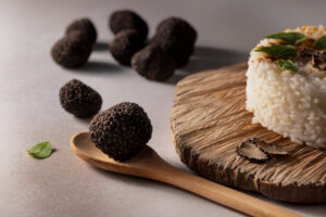 Black truffles resting on a wooden spoon with a mound of white rice on a rustic cutting board, garnished with greens and mushrooms.