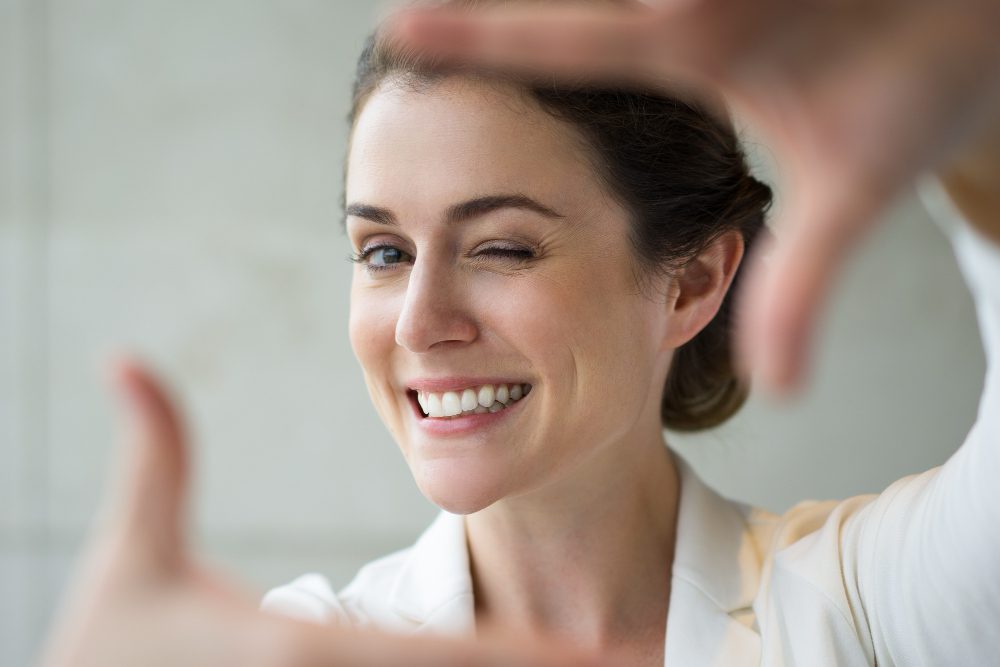 Smiling woman winking at the camera, hands forming a frame around her face as a photo gesture