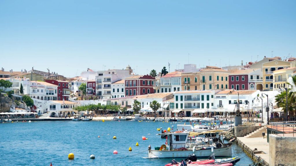 Harbor in a sunny Mediterranean town with colorful beachfront buildings and moored boats by the quay.