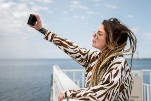 Person with long blond dreadlocks and sunglasses, wearing a zebra-pattern shirt, taking a selfie with a smartphone on a seaside boardwalk.