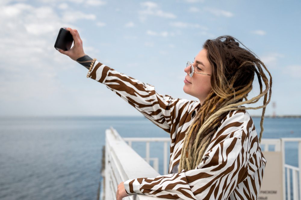 Person with long blond dreadlocks and sunglasses, wearing a zebra-pattern shirt, taking a selfie with a smartphone on a seaside boardwalk.